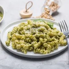 Pyszny makaron z pesto ze słoika, posypany parmezanem. Obok miseczka z zielonym sosem, kawałki sera i orzeszki piniowe.