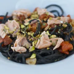 Czarny makaron z łososiem, pomidorkami i pistacjami na białym talerzu.