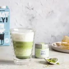 Warstwowy napój matcha z mlekiem owsianym, obok puszka z matchą i bambusowa miotełka. Idealny matcha przepis.