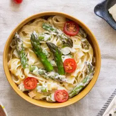 Pyszny makaron ze szparagami w sosie śmietanowym, udekorowany pomidorkami koktajlowymi. Obok miseczka z parmezanem i tarką.