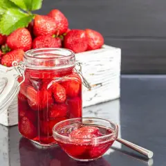 Słoik pełen domowego kompotu z truskawek, obok miseczka z owocami i syropem. Idealny sposób, jak zrobić kompot z truskawek na lato.