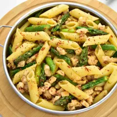 Penne z kurczakiem i szparagami, posypane płatkami chili, podane w metalowej misce na drewnianej desce.