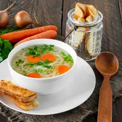 Aromaticzny rosół z perliczki z makaronem i marchewką, podany z grzankami. Obok leżą cebule i natka pietruszki.