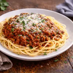 Pyszny sos do makaronu bolognese, obficie polany startym parmezanem i posypany natką pietruszki, na białym talerzu.