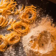 Świeży, domowy makaron, przygotowany według przepisu babci. Na ciemnym blacie leżą zwinięte w kółka tagliatelle i kopczyk mąki.