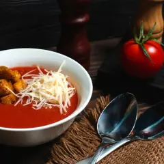 Aksamitna zupa pomidorowa z pomidorów z puszki, udekorowana grzankami i startym serem. Obok leżą łyżki i świeże pomidory.