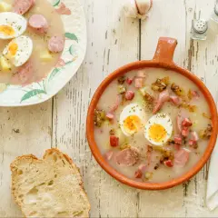 Dwa talerze z barszczem białym i żurkiem, pełne kiełbasy, jajek i ziemniaków, obok kromka chleba.