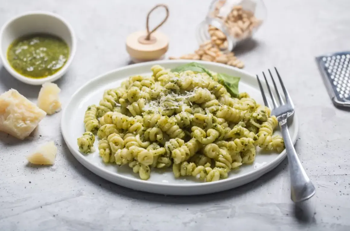 Pyszny makaron z pesto ze słoika, posypany parmezanem. Obok miseczka z zielonym sosem, kawałki sera i orzeszki piniowe.
