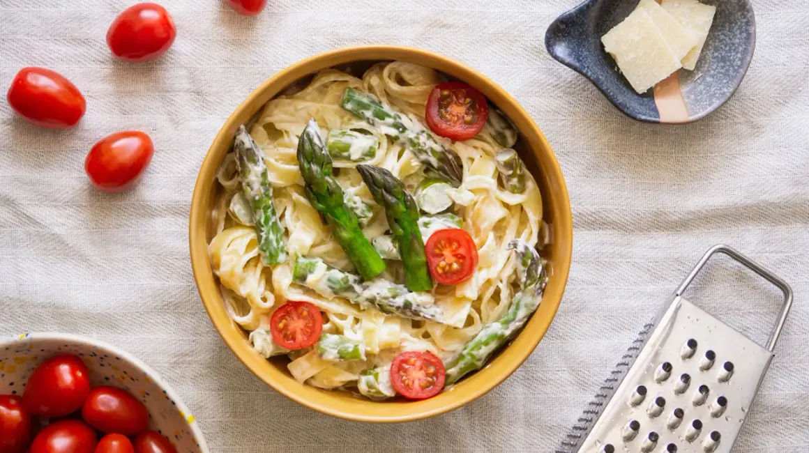 Pyszny makaron ze szparagami w sosie śmietanowym, udekorowany pomidorkami koktajlowymi. Obok miseczka z parmezanem i tarką.