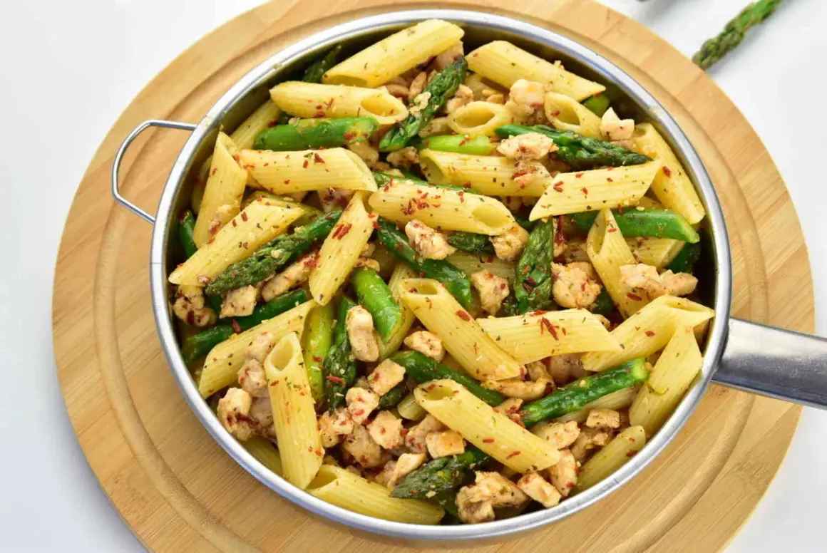 Penne z kurczakiem i szparagami, posypane płatkami chili, podane w metalowej misce na drewnianej desce.