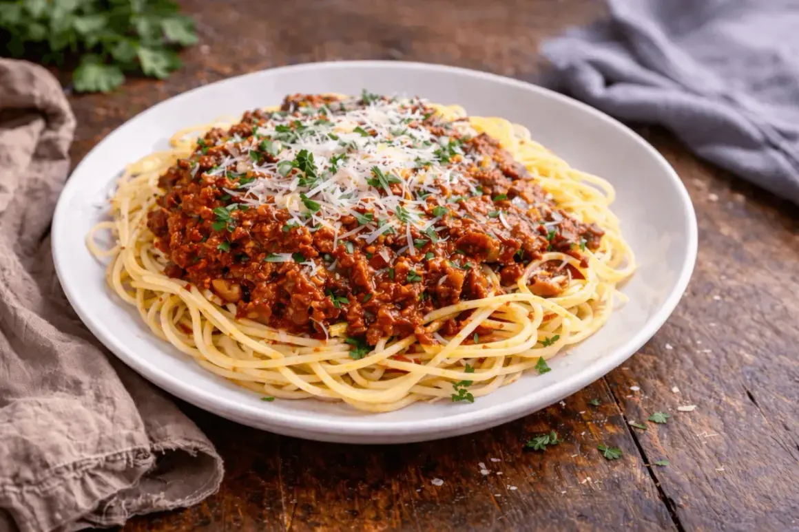 Pyszny sos do makaronu bolognese, obficie polany startym parmezanem i posypany natką pietruszki, na białym talerzu.