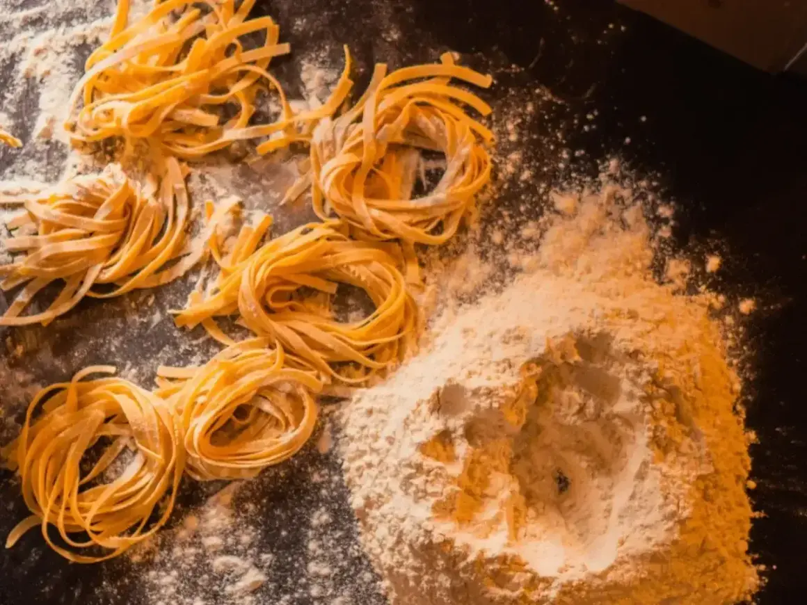 Świeży, domowy makaron, przygotowany według przepisu babci. Na ciemnym blacie leżą zwinięte w kółka tagliatelle i kopczyk mąki.
