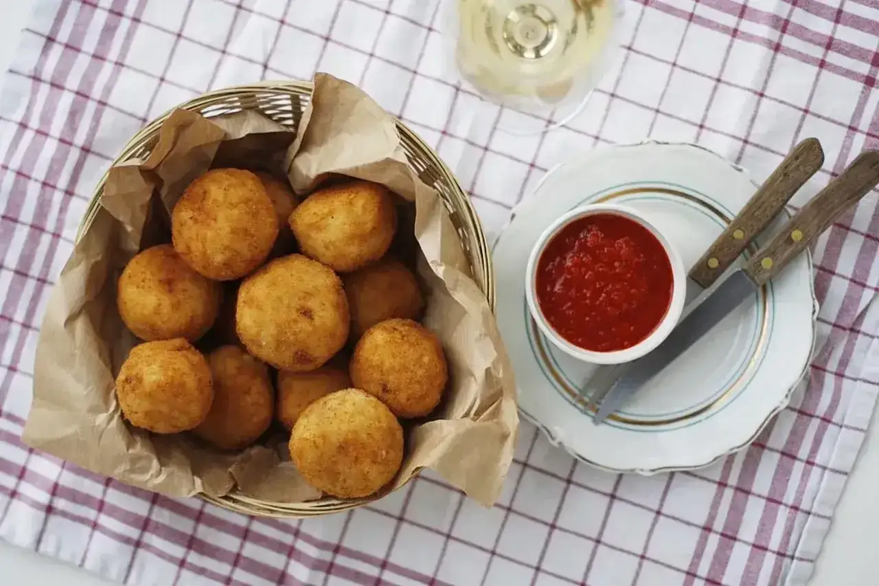 Koszyk pełen złocistych arancini, czyli smażonych kulek ryżowych, obok miseczki z sosem pomidorowym.